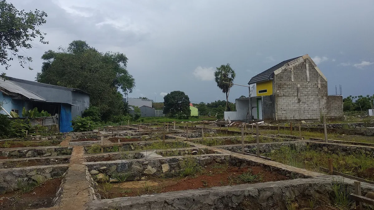 Pondasi batu untuk beberapa kavling rumah di lahan berumput, dengan satu rumah berdinding batako yang belum diplester dan sudah beratap di latar belakang